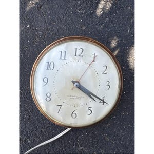 May include: A round Westclox electric wall clock with a metallic gold-toned frame and a cream-colored face. The clock features black Arabic numerals, black hour and minute hands, a thin red second hand, and a white power cord extending from the base.