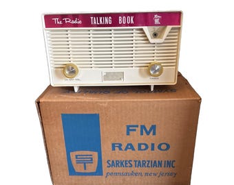Può includere: Una radio color crema etichettata The Radio TALKING BOOK con due manopole dorate, appoggiata su una scatola di cartone marrone stampata con FM RADIO SARKES TARZIAN INC e la località pennsauken, new jersey, con un logo blu con le lettere ST.