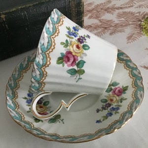 May include: A white teacup and saucer with a floral pattern in shades of pink, yellow, and blue. The cup and saucer have a gold trim and a turquoise border.