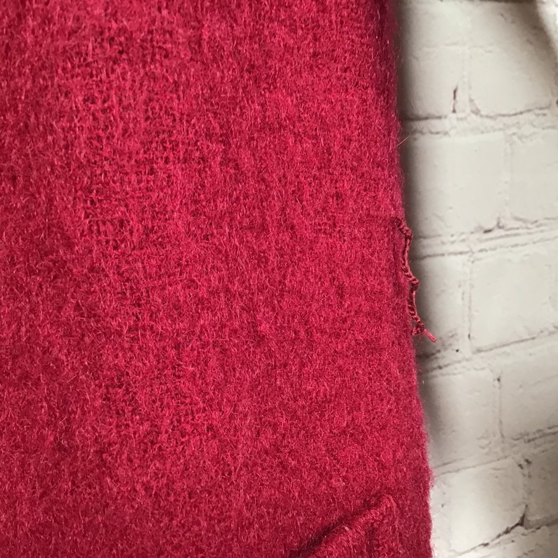Puede incluir: Primer plano de un su&eacute;ter rojo oscuro y peludo con un bolsillo en la parte inferior. El su&eacute;ter tiene un borde deshilachado en el lado derecho.