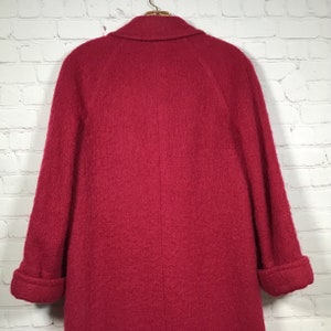 Puede incluir: Un abrigo largo de lana roja con cuello y mangas largas. El abrigo tiene una silueta ligeramente entallada y una superficie lisa y texturizada.