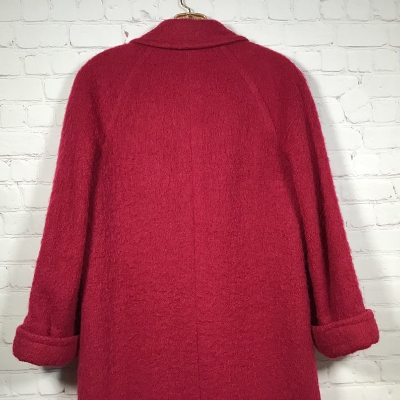 Puede incluir: Un abrigo largo de lana roja con cuello y mangas largas. El abrigo tiene una silueta ligeramente entallada y una superficie lisa y texturizada.