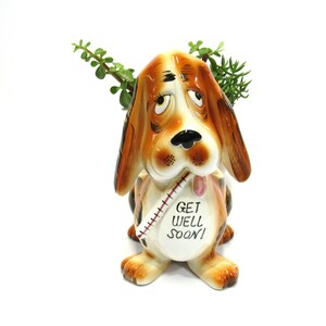 May include: A ceramic planter shaped like a dog with a thermometer in its mouth. The dog has brown and white fur and a sad expression. The planter says "Get Well Soon!" on its side.