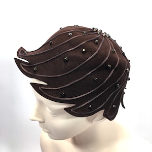 May include: A brown felt hat with a unique design featuring curved lines and black bead embellishments.