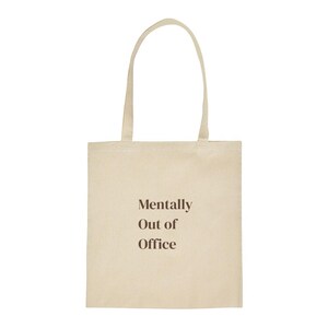 Könnte beinhalten: Eine beigefarbene Tragetasche aus Segeltuch mit zwei langen Schulterriemen. Auf der Vorderseite der Tasche befindet sich der Text "Mentally Out of Office", der in einer dunkelbraunen serifenlosen Schrift auf den Stoff gedruckt ist.