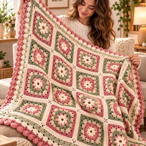 May include: A handmade crochet blanket featuring a repeating granny square pattern with floral motifs in shades of cream, sage green, and dusty rose. The blanket is finished with a scalloped border in a matching dusty rose yarn color.