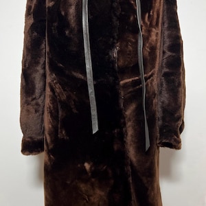 Peut inclure: Un long manteau marron foncé à la texture épaisse et pelucheuse. Le manteau présente un col montant et deux longs liens fins en similicuir marron foncé qui pendent de l'encolure. Le vêtement a des manches longues et une coupe droite.