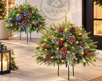 May include: Two spherical artificial evergreen shrubs on black metal stands, decorated with brown pinecones, red berries, and glowing lights. The front shrub features warm white lights, while the back shrub has multicolored lights.