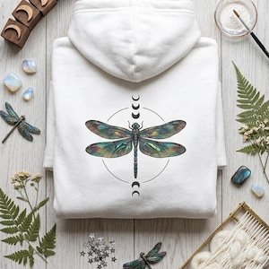 May include: A white hooded sweatshirt featuring a central graphic of a large, iridescent dragonfly with detailed wings, enclosed in a thin black circle with moon phase symbols above and below the insect. The design is printed on the back of the hoodie.
