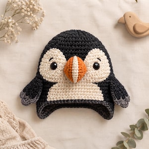 Puede incluir: Un gorro de ganchillo con diseño de frailecillo, que presenta un cuerpo gris carbón oscuro, cara y pecho de color crema, y un pico prominente de color naranja y blanco. El gorro tiene dos ojos negros y pequeñas solapas laterales grises.
