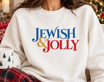 May include: A cream-colored crewneck sweatshirt featuring the text JEWISH & JOLLY printed on the front. The word JEWISH is in blue, the ampersand is in gold, and the word JOLLY is in red. The sweatshirt has long sleeves and a ribbed collar.