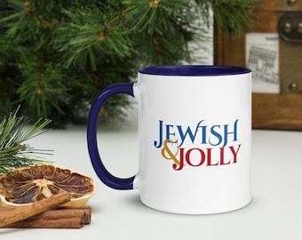 May include: A white ceramic mug with a dark blue handle and interior rim. The front of the mug features the text "JEWISH & JOLLY" printed in a stylized blue and red font, with the ampersand rendered in a gold color.