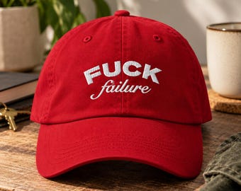 May include: A bright red baseball cap featuring white embroidered text on the front panel. The word FUCK is stitched in a bold, block-style font, while the word failure is stitched below it in a smaller, cursive script font.