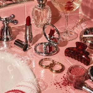 Puede incluir: Una colección de objetos que incluye un pintalabios rojo, un espejo de bolsillo plateado, pendientes de aro dorados, una pinza para el pelo de carey, un frasco de perfume de cristal, una copa de cóctel con una cereza y una brocha de maquillaje.