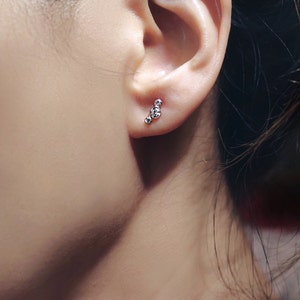 Pu&ograve; includere: Primo piano di un orecchio con un piccolo orecchino a perno d'argento con tre piccole pietre rotonde e trasparenti.