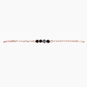 K&ouml;nnte beinhalten: Ein zartes Armband aus einer goldenen Kette mit vier kleinen runden schwarzen Steinen und einem wei&szlig;en Stein in der Mitte.