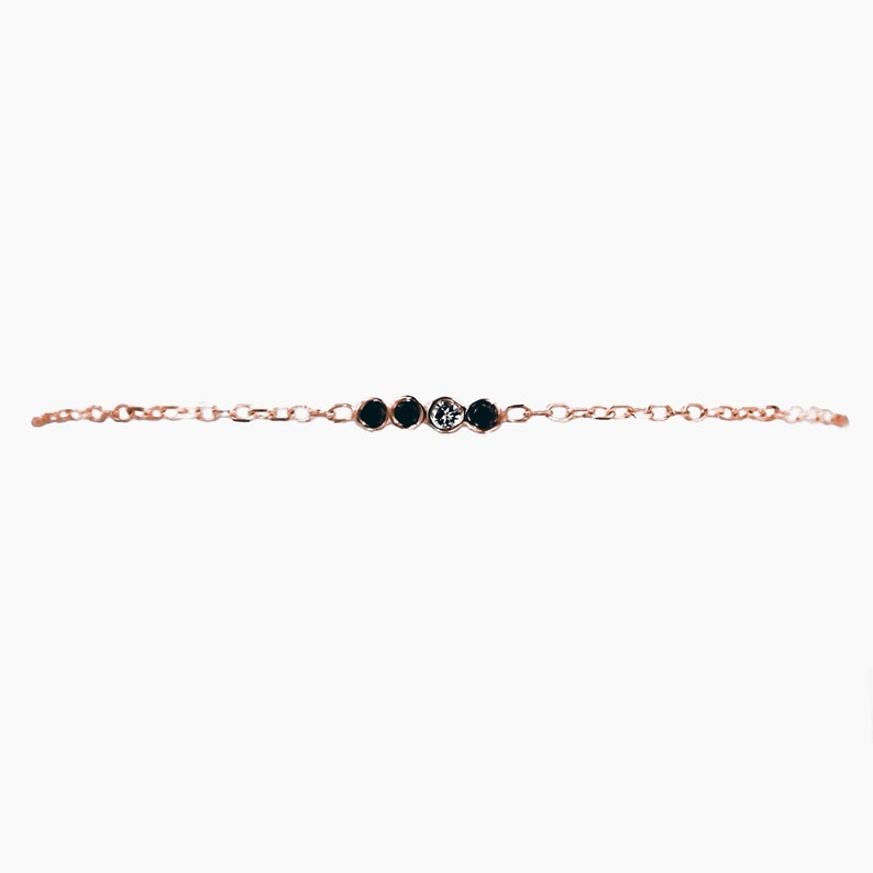 K&ouml;nnte beinhalten: Ein zartes Armband aus einer goldenen Kette mit vier kleinen runden schwarzen Steinen und einem wei&szlig;en Stein in der Mitte.