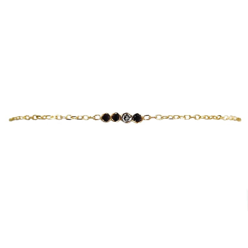 K&ouml;nnte beinhalten: Ein zartes Armband aus einer goldenen Kette mit f&uuml;nf kleinen runden Steinen. Die Steine sind schwarz und wei&szlig; und bilden ein schlichtes und elegantes Design.