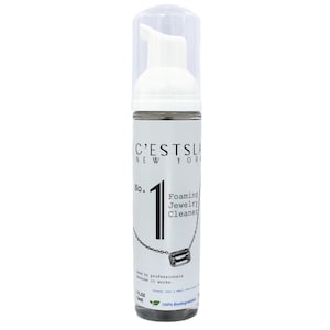May include: A white bottle of C'ESTSLA New York No. 1 Foaming Jewelry Cleaner. The bottle has a clear pump top and a label with text and a graphic of a necklace. The label also states the product is 100% biodegradable.