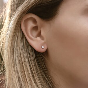 May include: A close-up of a person's ear with a small, square-shaped diamond stud earring.