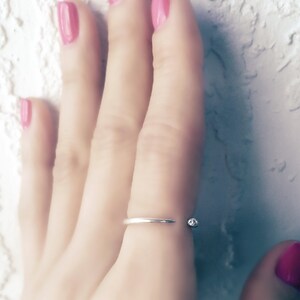 Op de afbeelding: Een zilveren ring met een kleine, heldere steen aan de zijkant. De ring wordt gedragen aan de middelvinger van een hand met roze nagellak.