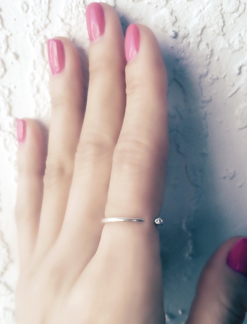 Op de afbeelding: Een zilveren ring met een kleine, heldere steen aan de zijkant. De ring wordt gedragen aan de middelvinger van een hand met roze nagellak.