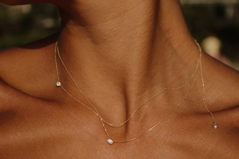 May include: A gold chain necklace with three small diamond pendants. The necklace is layered and features a round diamond pendant, a rectangular diamond pendant, and a smaller round diamond pendant.
