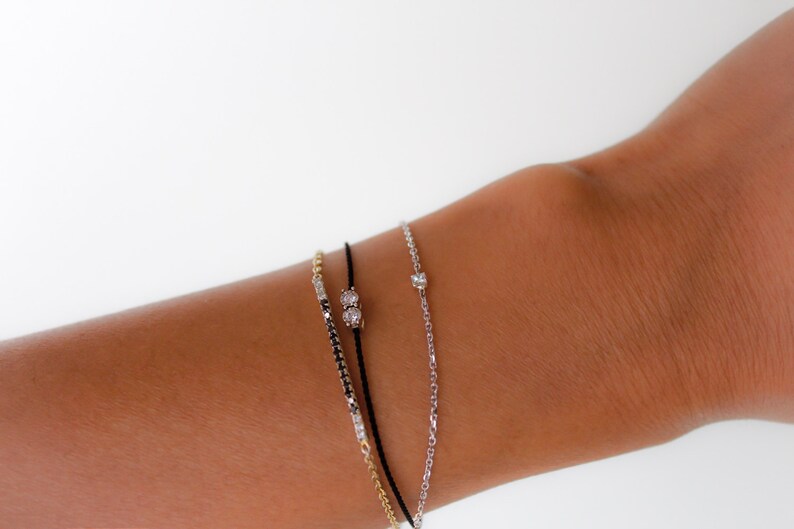 May include: Three bracelets on a wrist. The bracelets are a black string bracelet with a small diamond accent, a gold chain bracelet with a small diamond accent, and a silver chain bracelet with a small diamond accent.