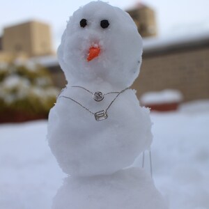May include: A snowman wearing two silver necklaces with diamond pendants. The snowman is made of snow and has a carrot nose. The background is blurry and out of focus.