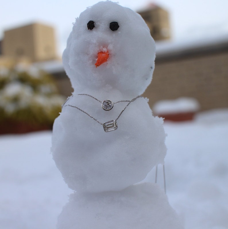 May include: A snowman wearing two silver necklaces with diamond pendants. The snowman is made of snow and has a carrot nose. The background is blurry and out of focus.
