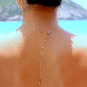 May include: A person with tanned skin wearing a delicate silver chain necklace with a small pendant. The necklace is long and hangs down the back.