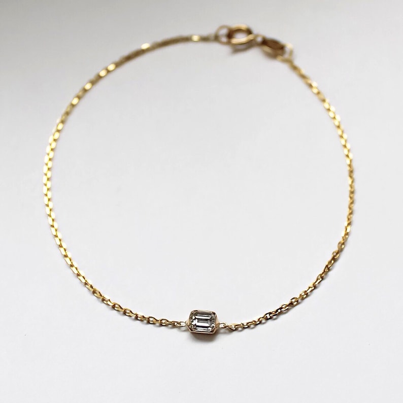 K&ouml;nnte beinhalten: Ein zartes Armband aus einer goldenen Kette mit einem einzigen, klaren, rechteckigen Edelstein.
