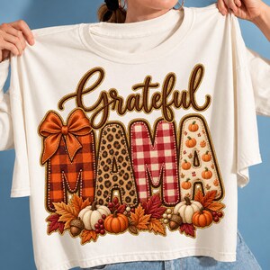 Peut inclure: Un t-shirt crème portant le mot Grateful en écriture cursive brune au-dessus du mot Mama. Les lettres sont remplies de motifs à carreaux orange, léopard et vichy rouge, agrémentées d'un nœud orange et de citrouilles d'automne.