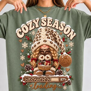 Peut inclure: Un t-shirt vert sauge avec un motif de gnome aux tresses blondes portant un bonnet à imprimé léopard. Le texte indique COZY SEASON et loading 87%. Le design inclut des branches de pin, des pommes de pin, des baies rouges et des flocons.
