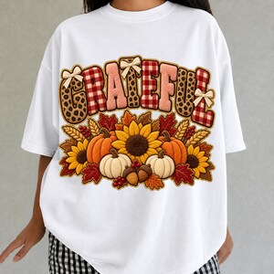 Peut inclure: Un t-shirt blanc avec le mot GRATEFUL écrit dans un mélange de motifs léopard et carreaux rouges, agrémenté de petits nœuds. Sous le texte, un motif de citrouilles orange et blanches, tournesols jaunes, feuilles d'automne et glands.
