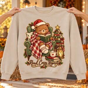Peut inclure: Un sweat-shirt beige à col rond orné d'un ours en peluche marron portant un bonnet de Père Noël, assis dans un fauteuil vert et lisant un livre sous une couverture à carreaux rouges et blancs. À côté se trouvent des livres, une lanterne et une tasse avec le texte Read, Cozy, Repeat.