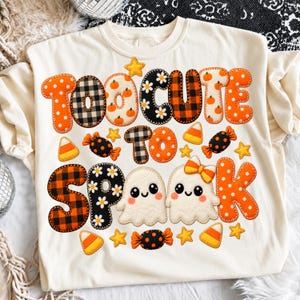 May include: A cream-coloured t-shirt featuring the text TOO CUTE TO SPOOK in large, patterned letters. The design includes orange polka dots, black and white plaid, and floral prints, alongside two small white ghosts and candy corn graphics.