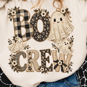 May include: A cream-coloured sweatshirt featuring the text BOO CREW. The letters are decorated with black and white buffalo plaid, floral patterns, and small embroidered flowers. Three ghost figures with eyelet lace details and bows are integrated into the design.