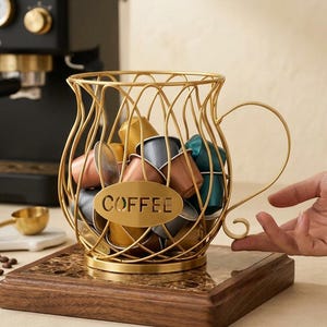 Könnte beinhalten: Ein goldfarbener Kaffeekapselhalter aus Draht in Form einer großen Tasse mit Henkel. Auf der Vorderseite befindet sich eine ovale Plakette mit dem ausgeschnittenen Wort COFFEE. Der Halter ist mit verschiedenen metallischen Kaffeekapseln in Gold, Kupfer, Blau und Grau gefüllt.