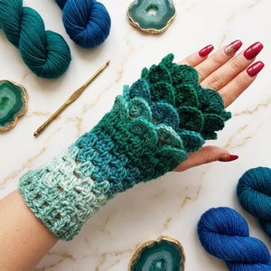 May include: A fingerless glove featuring a crochet scale pattern in an ombre gradient of light mint green to deep forest green. The texture consists of overlapping circular stitches that resemble dragon scales, extending from the wrist to the knuckles.