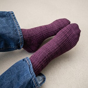 May include: A pair of dark purple knitted socks with a vertical ribbed texture. The socks are worn with blue denim jeans, showing the lower leg and foot area. The knit pattern features consistent vertical lines running from the cuff to the toe.