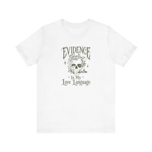 May include: A white short-sleeve t-shirt featuring a dark green graphic of a skull adorned with flowers and a butterfly. The text above the skull reads Evidence and the text below reads Is My Love Language, accompanied by small star icons.