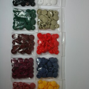 May include: Assortment of plastic snap buttons in various colors, including black, white, green, red, burgundy, blue, and yellow. The buttons are packaged in individual clear plastic bags.