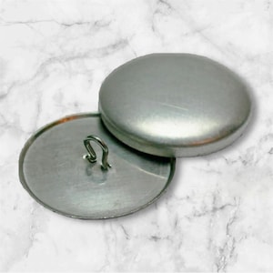 May include: A silver metal button with a loop on the back. The button is round and has a smooth surface.