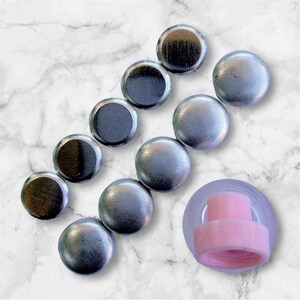 May include: Ten silver-colored round bottle stoppers with a smooth, slightly textured surface. The stoppers are arranged in a row on a white marble surface. A pink plastic bottle stopper is also visible in the image.