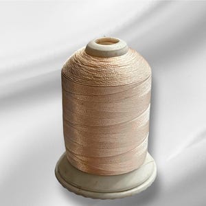 Peut inclure: Une bobine de fil de couleur pêche clair enroulée sur une bobine en plastique blanche.