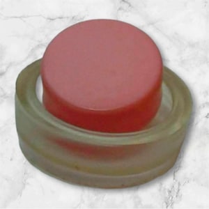 Puede incluir: Un tarro redondo rosa con una tapa de plástico transparente. La tapa tiene un borde elevado alrededor de la parte superior.