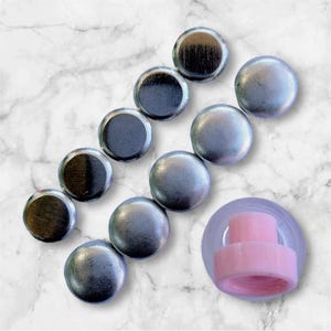 Może przedstawiać: Dziesięć srebrnych, okrągłych korków do butelek o gładkiej, lekko fakturowanej powierzchni. Korki ułożone są w rzędzie na białym marmurowym tle. Na zdjęciu widoczny jest również różowy plastikowy korek do butelek.