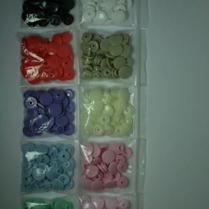May include: Assortment of plastic snap buttons in various colors, including black, white, red, purple, light blue, green, and pink. The buttons are packaged in clear plastic bags.