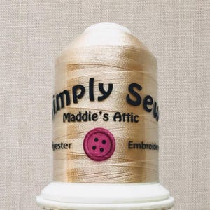 Peut inclure: Une bobine de fil à broder beige avec le texte "Simply Sew Maddie's Attic" et un bouton rose à quatre trous.
